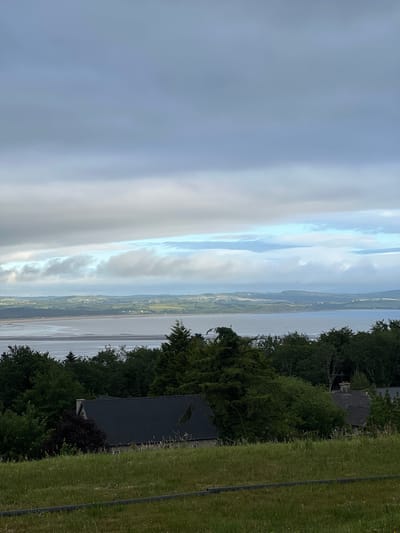 The image depicts a serene landscape from Mountcharles, Ireland, likely captured during the late afternoon or early evening, given the soft light. The scene is dominated by a vast expanse of water, possibly a bay or estuary, which occupies the central and left portions of the frame. Wispy clouds streak across a pale sky, creating a sense of depth and atmosphere.
To the right of the water, a lush green landscape unfolds, characterized by rolling hills and scattered vegetation. A dense line of trees forms a natural border between the water and the land. Within this treeline, hints of human habitation appear, with the dark roofs of houses peeking through the foliage. One house, in particular, is partially visible, featuring a dark roof and a chimney. The foreground to the far right is a neatly mown grassy area, suggesting a well-maintained garden or park. The overall mood is tranquil and picturesque, highlighting the natural beauty of the Irish coast. No visible text is present in the image.