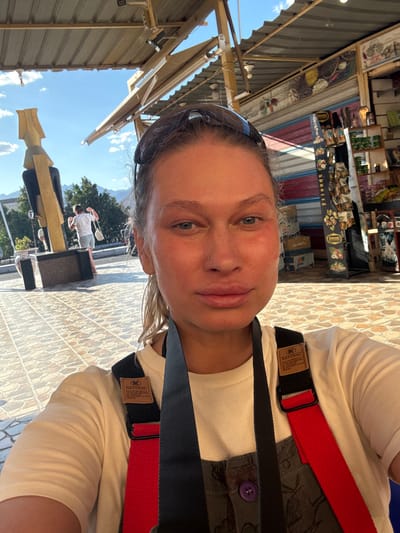 A selfie captures a woman in Dahab, Egypt, wearing a white T-shirt and a red and black harness. Behind her is a shop displaying tourist souvenirs, suggesting a market or tourist area. In the background, there's an individual walking on a tiled surface. The sky is clear blue, indicating a sunny day.