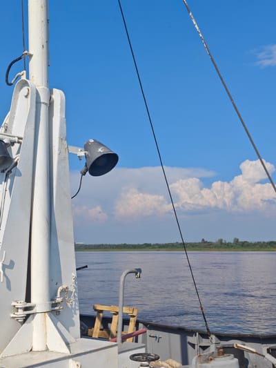 The image shows a view from a vessel on a body of water. In the foreground, a portion of the vessel's deck and superstructure is visible, primarily featuring a white metal mast assembly with attached fixtures, including a dark gray spotlight with a coiled cable. Another dark cable runs diagonally upwards across the frame, disappearing towards the top. The water is a deep blue with some ripples, and a distant tree-lined shore is visible on the horizon. The sky is a bright blue with scattered white cumulus clouds. The scene appears to be set during daylight with clear weather conditions. The location is identified as Verkhne-Pashino, Russia.