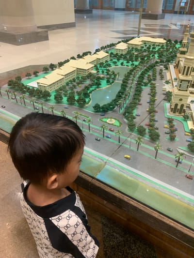 A young child, viewed from above and behind, is engrossed in observing a large, detailed architectural scale model displayed within an indoor setting in Samarinda, Indonesia. The child has dark hair and is wearing a white short-sleeved shirt patterned with a repeating black interlocking symbol, possibly a stylized 'C' or 'G', and black trim.

The scale model meticulously depicts an extensive development, featuring numerous light-colored, multi-story buildings. The landscape is rich with miniature green trees and shrubs, arranged along roadsides, within courtyards, and around a central body of water resembling a pond or small lake. The model includes a network of miniature roads with painted markings, several parking lots filled with tiny cars of various colors (yellow, red, blue), and numerous palm trees. A small circular green area with a central feature, possibly a fountain or roundabout, is also visible. Notably, text reading "SLAMET RIYADI" is faintly visible on one of the miniature roads within the model, likely indicating a street name or project title relevant to Samarinda.

The model is enclosed within a display case, with a glass barrier and a dark wooden frame separating it from the observer. The floor where the child stands appears to be light-colored tile. The scene suggests a moment of childlike wonder and engagement with the miniature representation of a built environment.