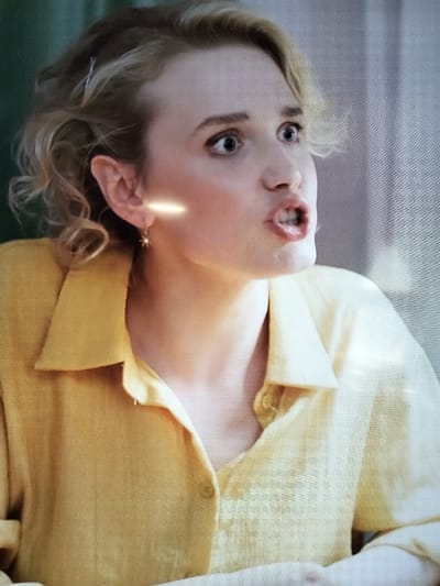 The media file shows a close-up, eye-level shot of a young woman from the shoulders up. She has blonde, curly hair pulled back loosely with a silver clip and a small star-shaped earring in her left ear. Her eyes are wide open and looking off to the side, and her mouth is open as if she is speaking or exclaiming. She is wearing a yellow collared shirt with buttons down the front. The background is blurred, with a dark green on the left and a textured, light-colored surface on the right, possibly a wall or curtain. A bright horizontal light streak is visible on her neck, near her ear, which appears to be a reflection or lighting effect. The overall impression is of a candid moment, possibly from a film or television show, capturing a moment of surprise or strong emotion. The context of Sloboda, Russia is noted, but no specific visual cues in the image confirm this location. The time of day and weather are not discernible. There is no visible text in the image.