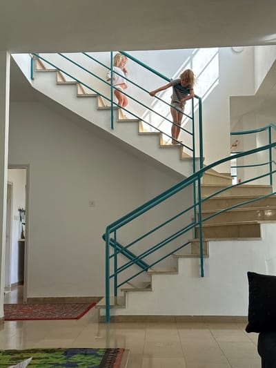 An interior scene in Kfar Vradim, Israel, depicts two young children on a multi-level staircase with a teal metal railing.
The environment consists of white walls, light tan/cream tiled flooring, and matching marble-effect stair treads with white risers. The prominent railing system is constructed from teal-colored metal, featuring a top handrail and multiple horizontal bars.
On the upper section of the staircase, a younger child with light blonde hair, wearing a white long-sleeved top and white underwear/diaper, stands facing right, holding the teal railing with both hands. Below and to the right of this child, a slightly older child with light blonde hair, wearing a striped short-sleeved top and white underwear/diaper, stands on a lower step, leaning forward and holding the railing with their left hand. Both children appear to be observing something below them or engaging with the stairs.
Strong natural light enters from an unseen source above and to the right, casting distinct diagonal shadows of the railing onto the white wall behind the children.
In the left foreground, a red patterned rug is partially visible on the tiled floor, leading to an open doorway revealing a glimpse of an interior hallway with objects on a wall fixture and a shelf. Another multicolored rug is partially visible in the bottom-left corner. On the white wall to the left of the staircase, an electrical outlet or switch is visible. A dark pillow is partially visible in the bottom-right foreground.