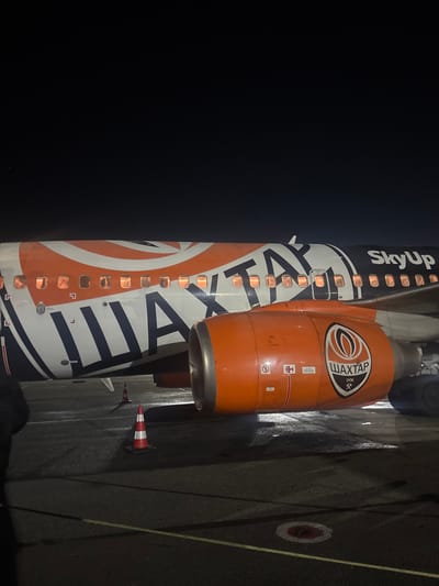 The image displays the mid-fuselage and right-side engine of a passenger aircraft at night on an airfield tarmac in Bubuieci, Moldova. The aircraft livery features a distinctive orange and dark blue/black pattern. The Cyrillic word "ШАХТАР" (Shakhtar) is prominently displayed in large white letters with black outlining across the orange section of the fuselage. The airline name "SkyUp" is visible in white near the upper aft section of the fuselage. The aircraft's engine nacelle is solid orange, featuring the logo of FC Shakhtar Donetsk, which includes "ШАХТАР 1936". Multiple passenger windows along the fuselage are illuminated from within. The tarmac below is dark, appearing wet with reflections of light. Two red and white striped traffic cones are positioned on the tarmac near the aircraft, with one partially obscured. A circular ground marking is visible on the tarmac. A partial view of a dark-clad individual is present on the far left edge of the frame, near the aircraft.