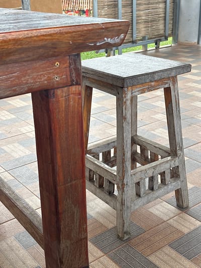 The scene is set in a semi-outdoor patio area in Ban Mai Saha Sampan, Thailand, under daylight conditions. The foreground features two weathered wooden furniture pieces. On the left, a reddish-brown wooden table with a rectangular top and robust square legs displays visible screws securing its structure. The table's top edge exhibits several white, irregular marks. To the right, a gray-toned wooden stool with a square seat and four legs stands adjacent. This stool includes a lower rectangular frame constructed from horizontal and vertical wooden slats, serving as a footrest or brace, with visible screws in its construction. Both the table and stool show signs of significant weathering, with faded finishes and surface wear. The floor beneath the furniture consists of multi-toned rectangular ceramic tiles arranged in a staggered or herringbone pattern, incorporating shades of brown, gray, and beige. The background, out of focus, reveals a metal railing or frame supporting horizontal bamboo or wooden screen elements, with green foliage visible beyond. No individuals are present in the image, and no active events are occurring.