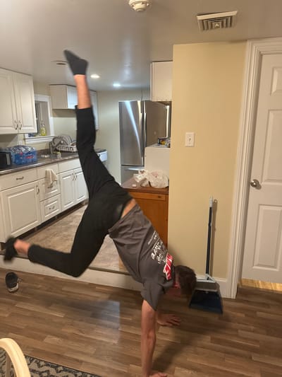 A person is performing a handstand in a kitchen in Ocean City, Maryland, United States of America. The person is wearing black socks, black pants, and a grey t-shirt with red lettering. Their left leg is extended upwards, and their right leg is bent. Their hands are on the floor, supporting their body. The kitchen features white cabinets, a stainless steel refrigerator, and a wooden floor. A broom and dustpan are standing in the corner next to a closed door. The lighting suggests it is daytime or the kitchen lights are on.