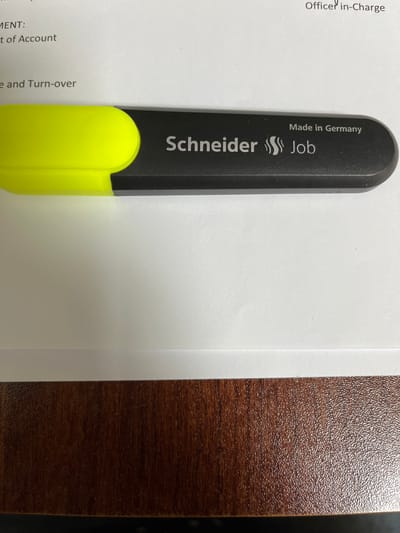 A close-up, high-angle shot shows a black and yellow highlighter pen positioned diagonally across a white document. The pen is positioned from the bottom left to the upper right of the frame. The yellow portion of the highlighter is on the left, and the black barrel with branding is on the right. The text on the black barrel reads "Made in Germany" and "Schneider Job". The document beneath the pen has some printed text, partially obscured by the pen, including the words "MENT:", "t of Account", and "e and Turn-over". The bottom of the image shows the edge of a wooden surface, likely a desk, with a dark brown, textured finish. There is no indication of the location in Quezon City, Philippines.
