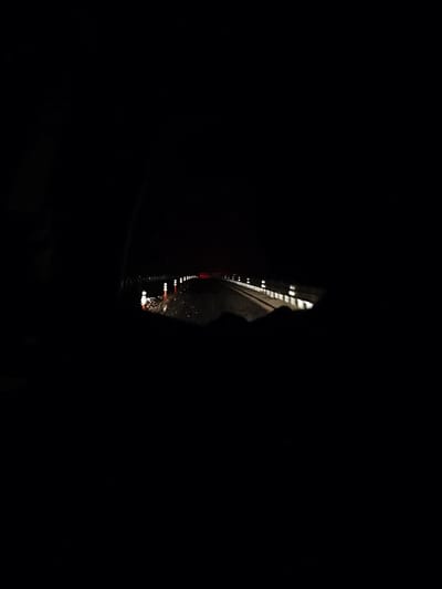 The image depicts a nighttime scene observed from inside a vehicle, located in Rashkovo, Bulgaria. The environment is characterized by deep darkness, with artificial light sources illuminating a wet road surface.
The primary subject is a paved road extending into the distance. On the left side of the road, approximately five vertical objects, consistent with road cones or bollards, are visible. These objects are white with reflective orange bands and are reflecting light, suggesting they are part of road-marking or safety infrastructure. The road surface itself is wet, creating elongated reflections of these light sources and other distant lights.
On the right side of the road, a barrier or guardrail is visible, also featuring multiple reflective markers or lights that recede into the distance. Farther along the road, a single, distinct red light is present, positioned centrally, which may indicate a distant vehicle's taillight or a signal.
The immediate foreground and the left periphery of the image are dominated by dark, out-of-focus shapes, consistent with the interior elements of a vehicle (e.g., dashboard, window pillar) from which the photograph was captured. No individuals or specific actions are discernible within the frame. The scene's illumination relies solely on these scattered artificial light points against the pervasive darkness.
