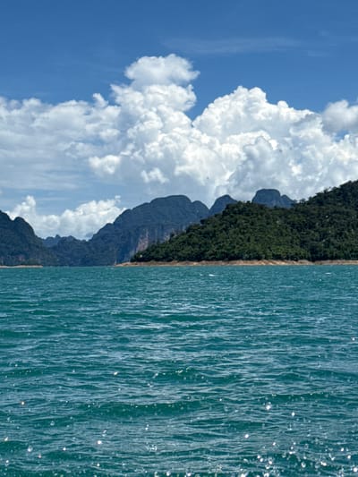 The image captures a scenic view of the Cheow Lan Lake in Khao Sok National Park, near Ban Chiao Lan, Thailand. The foreground features the tranquil, blue-green water of the lake, with small ripples and occasional glints of sunlight on its surface. In the middle ground, a lush green, tree-covered island rises from the water, with a sandy shoreline. Beyond the island, a series of dramatic, dark, and rugged karst mountains dominate the background, their peaks softened by distance and the haze. The sky above is a vibrant blue, dotted with large, fluffy white cumulus clouds, some of which cast subtle shadows on the landscape. The scene evokes a sense of natural beauty and serenity, typical of this region known for its stunning limestone formations and clear waters.