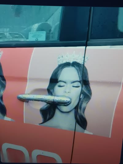 The image displays a close-up view of a vehicle door, likely in Serhiivka, Ukraine. A large graphic on the door features a portrait of a woman wearing a tiara, with her eyes closed and her mouth open as if to bite down on the car's door handle. The door handle itself is visible in the foreground, partially obscured by the graphic. The background of the graphic is a light pink color. The vehicle appears to be a light-colored car, with a portion of a window visible in the upper left corner, showing some rain droplets on the glass. The bottom of the image shows a portion of the car's body, with what appears to be part of a logo or text in a white outline on a pink background. The overall impression is a photograph taken from an oblique angle, possibly a snapshot of a vehicle encountered in the city.