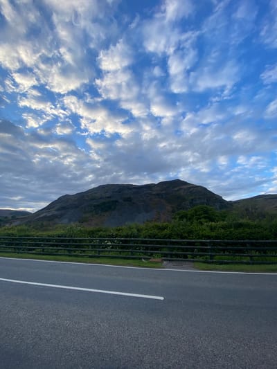 The image captures a scene in Llangynog, United Kingdom. The frame is divided into three primary sections. On the left, a cloudy blue sky fills the majority of the space. The sky is dynamic with varying shades of blue and white. Adjacent to the sky is a mountain partially covered in greenery, sloping downwards. A fence runs along the mountain. To the right of the fence is a paved road with a white lane marker. The road occupies the remaining portion of the image. The perspective is slightly angled, giving a view across the landscape. No people are visible.