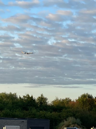 An airplane is captured mid-flight against a cloudy sky. Below the sky, a dense line of green trees stretches across the frame, with some autumnal colors visible in the foliage. In the foreground, the tops of some buildings are visible, one constructed of grey brick. The scene appears to be located in Kampenhout, Belgium, given the context provided.