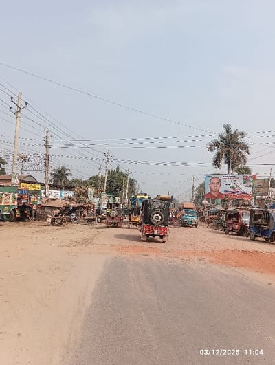 The image displays a daytime street scene in Kaliganj, Bangladesh, recorded on 03/12/2025 at 11:04. The view is of a wide, unpaved and asphalt road intersection or market area under a light blue sky crisscrossed by numerous overhead utility lines.
In the foreground, a red three-wheeled vehicle with a rear-mounted spare tire is positioned on the asphalt, facing away from the viewer. Multiple other three-wheeled auto-rickshaws, blue utility vehicles, and larger trucks, including one heavily decorated with green and blue accents, are distributed across the road. To the left, a cluster of informal market stalls or small structures lines the roadside.
On the right, a prominent palm tree is visible near a large billboard featuring a male portrait, likely a political figure. Utility poles are densely arranged, supporting extensive wiring. Human activity, primarily involving vehicle operation and presence near stalls, is observable. The ground consists of both paved sections and areas of reddish-brown dirt and gravel.