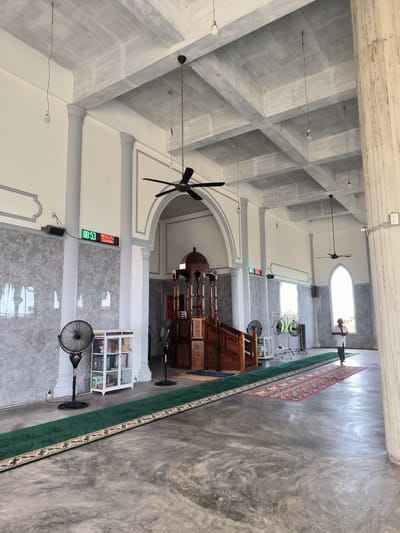 The image shows the interior of a mosque in Blangbitra, Indonesia. The mosque features a high ceiling with exposed concrete beams, supported by decorative pillars. Hanging from the ceiling are several ceiling fans and bare light bulbs. Along one wall is a digital clock displaying "00:53 MENUJU IQOMAH." Near the clock are a bookshelf, and several electric fans. A green carpet runner extends down the length of the room, leading to an elevated wooden minbar with stairs. Toward the back, a man in white is standing near an arched window. The floor is gray concrete.