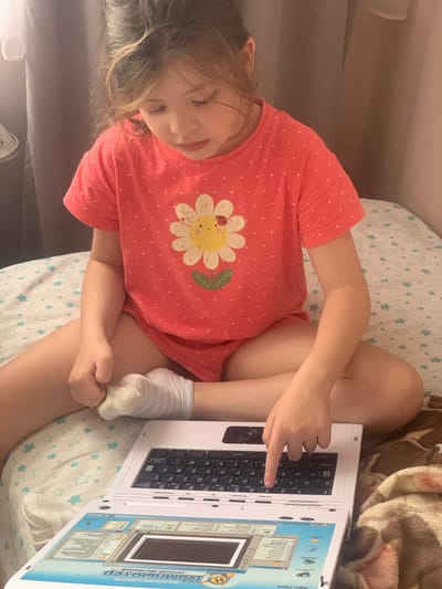 A young girl is lying on a bed, playing with a toy laptop. She is wearing a pink t-shirt with white polka dots and a flower design. The laptop is white and has a keyboard with Cyrillic letters. The screen displays what appears to be a children's educational program. The girl is looking down at the laptop with a relaxed expression. The scene is indoors, with a blanket draped over the bed and a pillow with a star pattern. The lighting suggests it could be daytime or early evening. The girl's position and the presence of the toy laptop indicate she is engaged in play or learning. The visible text on the laptop includes "KOMUGP" and "АВТСКИЙ ОБУЧ", which suggests it's a Russian educational toy laptop.