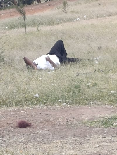 In the city of Matungulu west ward, Kenya, an outdoor scene depicts a single individual lying prone on their back in an expanse of dry, light green and yellowish grass. The person is dressed in a white top and a black garment that covers their lower body and appears to be pulled over their head, obscuring their face. Their arms are bent, with one hand visible near the chest area and the other arm extending to the right. One bare foot or lower leg is visible on the left side of the frame. The individual is positioned diagonally across the midground, with their head to the left and feet to the right.
In the immediate foreground, a reddish-brown, indistinct clump of material rests on a patch of bare dirt. The ground transitions from a dirt path in the foreground to the grassy area where the individual lies. Scattered white debris, possibly plastic bags or paper, is visible within the grass around the person. The background features more sparse grass, small trees or shrubs, and a dirt path or road. The overall environment appears to be an undeveloped or semi-urban outdoor area under natural daylight.