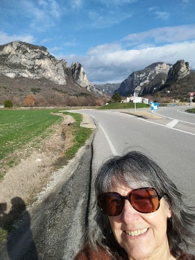 The image depicts an outdoor scene in Francillon-sur-Roubion, France, featuring an adult female in the foreground taking a selfie. The woman occupies the lower right portion of the frame, smiling with visible teeth, and wearing large, dark brown tortoiseshell sunglasses. Her gray hair is visible around her face, and a shadow on the lower left confirms a selfie being taken by a person holding a device.

The background shows a paved asphalt road curving to the right from the immediate foreground, with a solid white line marking its edge. A green grassy field, possibly agricultural, runs along the left side of the road, separated by a narrow dirt verge. In the middle distance, the road forms a roundabout or intersection marked by a red "Yield" sign and a blue "mandatory direction right" sign. Beyond the road and fields, prominent rocky mountains with steep, rugged faces dominate the horizon, particularly on the left and center. The mountainsides are sparsely covered with deciduous trees, appearing bare, alongside some evergreen vegetation. A few white buildings are visible at the base of the mountains near the road. The sky is partially cloudy with blue patches, and strong sunlight is evident, casting shadows.
