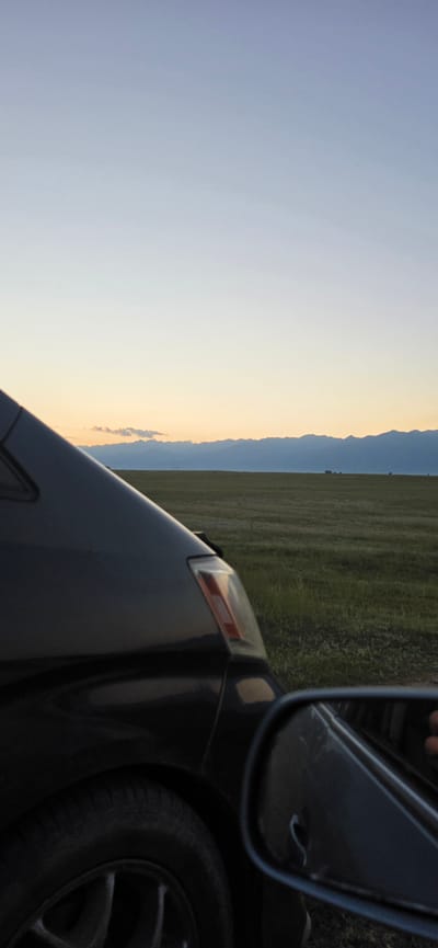 The image captures a scene from a vehicle in the Issyk-Kul region of Kyrgyzstan. The foreground is dominated by the dark exterior of a car, showing the front quarter panel, wheel, and part of a side mirror. The side mirror reflects a hand. Beyond the car, a vast, flat grassy field stretches towards a distant mountain range that forms a sharp silhouette against a gradient sky. The sky transitions from a pale blue at the top to a warm, soft yellow near the horizon, indicating either sunrise or sunset. The overall impression is one of a journey through a natural landscape at the edge of day.