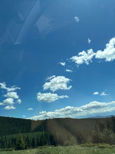 The image depicts a vast expanse of a clear blue sky dotted with scattered white clouds. In the upper portion of the sky, a circular, iridescent object, possibly a lens flare or a reflection, is visible. Below the sky, a dense forest of evergreen trees covers a hilly terrain, creating a dark green band across the middle of the frame. The forest appears to be on a mountainside, with the trees growing closely together. In the foreground, there is a grassy area with some low-lying bushes and scattered rocks. The overall scene suggests a natural, outdoor environment, likely in a mountainous or forested region. There are no people or specific man-made structures clearly visible that would pinpoint a location in Cherna Mesta, Bulgaria, but the general landscape is consistent with a rural or wilderness setting.
