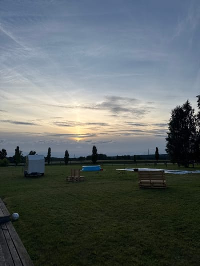 The image depicts an outdoor scene at dusk in Jeedasküla, Estonia. The sky is filled with wispy clouds and the sun is setting, casting a warm glow on the horizon. In the foreground, a well-maintained grassy field is visible, with a wooden deck on the left side. A white soccer ball rests on the deck. Scattered across the field are several objects, including a small white trailer, a blue inflatable pool, stacks of wooden pallets, and a wooden bench made from pallets. In the background, a line of trees borders the field, and beyond them, a rural landscape with fields and more trees can be seen. A power line pole is also present in the distance. The overall impression is one of a peaceful, recreational outdoor space at the end of the day.