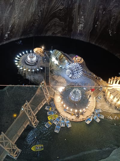 The image depicts a subterranean recreational area within the Turda Salt Mine in Turda, Romania. The scene is viewed from an elevated perspective, looking down into a large, cavernous space with dark, textured walls that show stratified rock formations.
In the center of the frame, several illuminated structures are built on a rocky platform emerging from a dark body of water. One structure features a circular wooden deck with numerous lights radiating outwards from a central point. Adjacent to this is a large, geodesic dome-like structure covered in lights. Further along the platform, a transparent, curved structure and another circular wooden deck with seating areas are visible. Wooden walkways and staircases connect these different areas.
Scattered on the dark water are several small boats, some yellow and some blue, most appearing to be unoccupied. One yellow boat in the foreground contains two individuals. Another small boat, with a single occupant, is visible further out in the water, closer to the cavern walls. A large net structure covers a significant portion of the lower part of the image, extending over the water and connecting to wooden walkways.
People are present on the walkways and platforms, interacting with the structures and the environment. The overall impression is of a well-lit, modern recreational space carved into an ancient geological formation. The lighting creates a dramatic contrast with the dark surroundings of the salt mine.