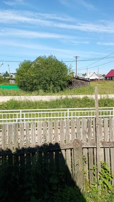 In Kochyovo, Russia, a sunny daytime scene unfolds, featuring a weathered wooden fence in the foreground. Behind the fence, a grassy area with scattered vegetation is visible, leading to a road or dirt path. Further back, a large, dense green bush dominates the middle ground, partially obscuring a pile of dark objects, possibly construction materials, and a small wooden shed with a lattice design. In the background, several houses with varying roof colors, including red and white, are scattered across the landscape under a bright blue sky with wispy white clouds. Power lines and utility poles are also present, indicating a residential area. The overall impression is a rural or semi-rural setting on a clear day.