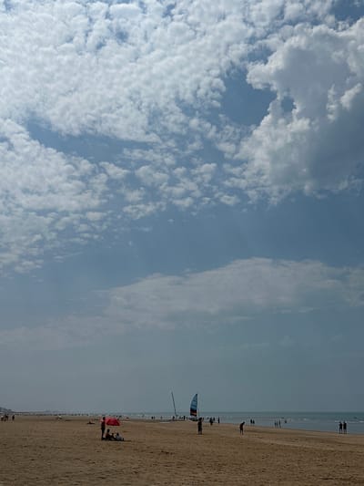This image depicts a beach scene in Koksijde, Belgium. The photograph is oriented vertically, with the sky occupying the left two-thirds and the beach and ocean filling the right third. The sky is a light blue, partly obscured by fluffy white clouds that are dense and textured in the upper left and become more wispy and diffused towards the right.

On the sandy beach, several people are visible. In the foreground, a person is lying on the sand. Further down the beach, a group of people are gathered under a bright red umbrella. A small sailboat with a blue and white sail is positioned on the sand near the water's edge, with a person standing beside it. In the distance, a long line of people can be seen walking along the shoreline, and the ocean stretches out to the horizon, appearing calm and blue.

The lighting suggests it is daytime, likely a bright day given the clarity of the sky and the contrast of the sand. The overall atmosphere appears relaxed and leisurely, typical of a beach day. The presence of the ocean and the sandy shore are clear indicators of a coastal location. No visible text is present in the image.