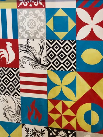 The image shows a grid of colorful squares, each with a distinct design. There's a lemon on blue, geometric patterns, a red and white flower design, and a black and white diamond pattern. Other squares feature stripes, stylized floral elements, a rooster silhouette, and a stylized red dolphin shape. The color palette includes red, blue, yellow, white, and black. The designs appear flat and graphic.