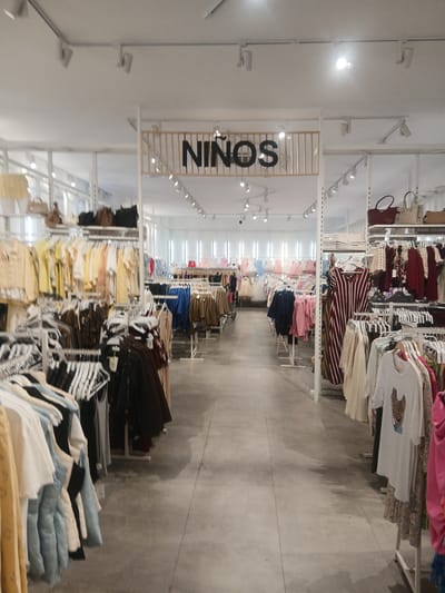 The image shows the interior of a clothing store, specifically the "Niños" (children's) section. The store appears well-lit with fluorescent lighting. Rows of clothing racks display a variety of children's clothes, including dresses, shirts, and jackets, in a range of colors. Some mannequins are visible, also dressed in children's attire. Bags are displayed on shelves above some of the clothing racks. The floor is concrete and the overall aesthetic is modern and clean. The location is identified as being in Ávila, Spain.