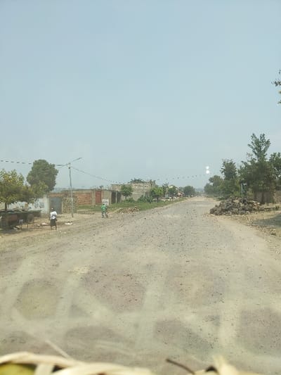 The image depicts an unpaved, dusty road traversing a semi-urban area within the city of Rutshuru, Democratic Republic of the Congo, under a pale blue, hazy sky. The perspective is slightly elevated, possibly from a vehicle, with blurred light-colored foliage visible in the immediate foreground.
The road, appearing uneven with visible ruts and scattered small stones, extends from the foreground into the distance. On the left side of the road, a dark-colored, two-wheeled flatbed trailer is parked. Adjacent to it, a person wearing a light-colored top and dark trousers stands facing away from the viewer. Further down the road on the left, several structures made of reddish-brown brick and unpainted concrete are visible, some appearing partially constructed or in disrepair. Utility poles with overhead wires extend along this side of the road.
In the center of the road, a person wearing a light green shirt and dark pants is riding a bicycle away from the viewer. On the right side of the road, a large pile of dark, irregularly shaped rocks, possibly volcanic material or construction debris, is present. Sparse green trees and other vegetation line both sides of the road, particularly towards the mid-ground and background. The distant background shows more scattered buildings and trees as the road recedes. Two distinct circular light anomalies are present in the upper-right portion of the sky.