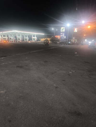 The image depicts a nighttime scene at a Pertamina gas station and a KFC restaurant in Meunasahceubrek, Indonesia. A bright, white illuminated canopy covers multiple gas pumps, with a vertical sign displaying "PERTAMINA" and fuel prices. Adjacent to it, a KFC sign featuring the iconic Colonel Sanders is visible. Several vehicles, including cars and motorcycles, are present on the asphalt road and within the gas station premises. A three-wheeled vehicle is parked near the gas station. The lighting from the gas station and streetlights creates a strong glare, and some vehicles' headlights are also illuminated. The ground is asphalt, with some scattered debris. The overall environment appears to be a busy intersection or commercial area at night.
