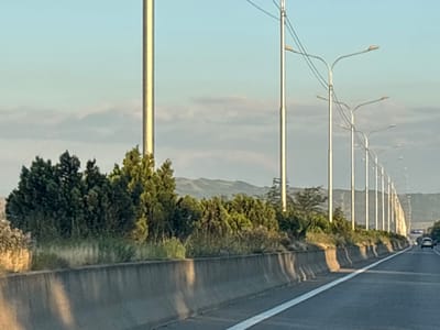 The image depicts a highway scene in Dzveli Kanda, Georgia. A concrete barrier lines the right side of the road, with a white line marking the edge of the travel lane. A line of tall streetlights with curved arms extends along the roadside, with one vehicle visible in the distance on the highway. Dense green trees and vegetation grow behind the barrier, with rolling hills in the background. The sky is a light blue with scattered clouds. The lighting suggests it is either early morning or late afternoon.