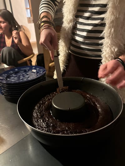 A person wearing a white fur vest and a black and white striped shirt is using a metal spatula to push a piece of chocolate cake in a black pan. The pan has a black ring in the middle. The cake is surrounded by a dark brown liquid. The person is standing over a stainless steel countertop. In the background, a stack of blue and white patterned plates sit on the counter. A blurred out person with brown hair is sitting in a wooden chair in the background. The scene takes place indoors in a kitchen. The coordinates provided place this location in Celerina/Schlarigna, Switzerland.