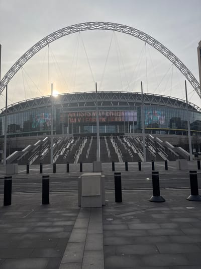 The image captures the exterior of Wembley Stadium in Wembley, United Kingdom, under bright, slightly hazy conditions, likely during early morning or late afternoon due to the low sun. The scene is dominated by the stadium's distinctive white arch, which spans gracefully across the sky above the main structure. The sun's glare is prominent behind the stadium's facade, particularly to the left of its central point, indicating its low position.
The stadium building features a modern design with large glass panels and a visible structural framework. On one of its exterior screens, faint white text is displayed. Partially legible phrases include "ARTISTS MAY ENTER" and fragments such as "IN REAL" and "UNIVERSE," possibly part of an advertisement or a general message.
In the foreground, a wide, paved plaza or concourse extends towards the stadium. This area is completely devoid of people, suggesting it is not an event day or that the image was taken during off-peak hours. Several black, cylindrical bollards are systematically arranged along the paved walkway, interspersed with square concrete plinths. Further back, metal barriers or queueing structures are visible on the steps leading up to the stadium entrance. The overall impression is one of architectural grandeur and scale, captured in a moment of quiet anticipation or dormancy.