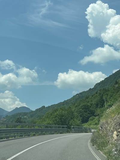 This is a photograph taken from a vehicle moving along a winding road in a mountainous region, likely in Ossimo Inferiore, Italy. The image is oriented vertically. On the right side of the frame, the asphalt road curves, with a white line marking its edge. A metal guardrail runs parallel to the road, separating it from a steep, heavily forested mountainside. The trees are a vibrant green, suggesting a summer or spring day. The sky is a clear blue, punctuated by several fluffy white clouds, some of which appear wispy. In the distance, parts of the mountain range are visible, with some peaks shrouded in shadow. The overall impression is one of a scenic drive through a natural landscape. The time of day appears to be daytime, with bright sunlight illuminating the scene. There is no text visible in the image.