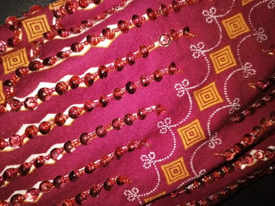 A close-up, angled forensic view captures a textile object in Na Sang, Laos. The primary material is a deep red or burgundy fabric patterned with repeating motifs. These motifs include yellow-orange squares containing concentric squares, and intricate white dotted lines forming curves and stylized floral or clover shapes. Several parallel, slightly wavy lines of translucent, reddish-orange sequins are stitched onto the fabric, extending diagonally from the upper-left to the lower-right of the frame. The sequins are disc-shaped, individually spaced, and exhibit light reflection variations. No individuals, actions, or specific environmental features beyond the fabric itself are discernible within the frame.