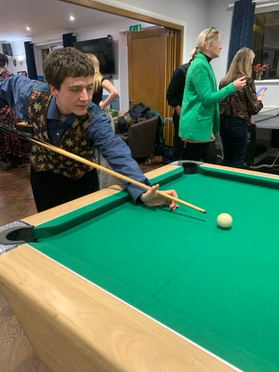 A young man in a patterned vest and blue shirt lines up a pool shot on a green felt table. He holds a cue stick, aiming at a white ball. Behind him, two women stand in a room with blue walls, a television, and various decorations. One woman wears a green blazer, while the other is in a patterned top and jeans, holding a phone. The scene appears to be an indoor gathering or party.
