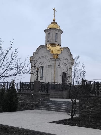 An exterior view depicts an Orthodox Christian chapel situated in Petrivske, Ukraine, under an overcast sky. The chapel features a light gray, textured stone facade with decorative arched recesses and vertical fluting. A prominent golden dome, capped with a smaller cylindrical drum containing vertical windows, supports a golden Orthodox cross. The structure stands on an elevated platform, accessible by a set of gray stone steps from the foreground. The platform is bordered by a dark, ornate wrought-iron fence. To the left of the chapel, on the platform, a dark stone pedestal holds an antique-style street lamp. The immediate foreground shows a light gray paved walkway with areas of dark soil. Bare tree branches, devoid of leaves, are visible on both the left and right sides, partially framing the chapel. No individuals are present in the scene.