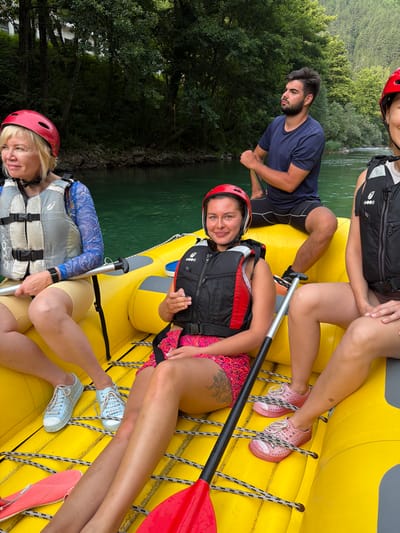 Four people are sitting in a yellow raft on a river. The raft is equipped with ropes and paddles. A man in a blue shirt is in the back, holding a paddle. Three women are in the front, wearing life jackets and helmets. The woman on the left is wearing a blue and white patterned shirt and beige shorts. The woman in the middle is wearing a red and black life jacket and pink patterned shorts. The woman on the right is wearing a black life jacket and dark shorts. The raft is on a green river, with a forested mountainside in the background. The location is Polje Bijela, Bosnia and Herzegovina.