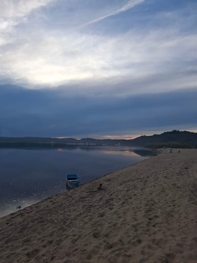 The image depicts a serene riverbank scene at dusk. The sky is filled with dramatic, layered clouds, ranging from deep blues and grays to lighter, diffused whites where the sun is setting. A vast, calm body of water reflects the muted colors of the sky and the distant, tree-covered hills that form the opposite bank. In the foreground, a sandy beach slopes down towards the water. A small, light blue rowboat is beached on the sand near the water's edge. Further down the beach, several small figures can be seen, suggesting a small gathering or individuals enjoying the evening. The overall impression is one of tranquility and the fading light of day. The location is identified as Malaya Sazanka, Russia.