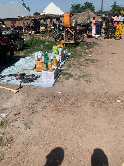 This image captures a busy outdoor market scene in Nigeria, likely during daylight hours under a clear sky. The foreground features a stall laid out on a patterned blue and white cloth, displaying an assortment of items. These include numerous bottles of various sizes and colors, many appearing to be cleaning products or beverages, alongside some cardboard boxes. Among the bottles, a box with the label "HORIZON OIL" is clearly visible, along with another box labeled "STYLE". Scattered on the cloth are also various tools, including what looks like a sickle and what appears to be a rifle or its components, suggesting a diverse range of goods for sale.
In the background, a collection of motorcycles is parked, indicating transportation is a significant aspect of the location. Behind the motorcycles, a makeshift structure with a thatched roof is visible, suggesting a semi-permanent market setup. Several people are present, some standing and interacting near the stall and motorcycles, while others are seen in the background, dressed in traditional Nigerian attire. The overall atmosphere appears lively, with the hustle and bustle typical of a local marketplace. The ground is a mixture of dirt and gravel, with sparse patches of grass, consistent with an outdoor setting in a rural or semi-urban area of Nigeria. The presence of motorcycles and the style of clothing suggest a contemporary setting.