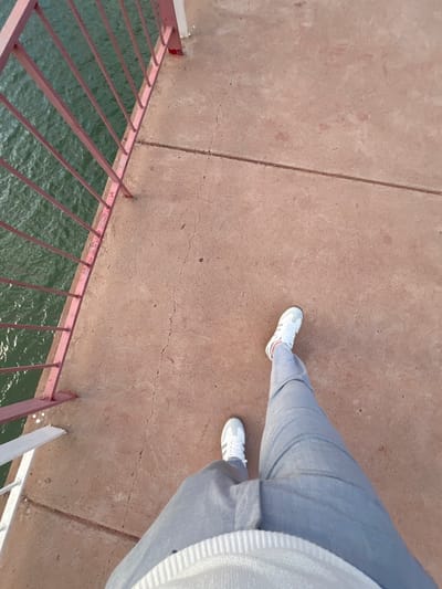 The image shows a downward-angled view from a bridge or elevated walkway in Tabaroucht, Morocco. The person standing on the structure is wearing light-grey trousers and white sneakers. The surface they are standing on is concrete, showing some cracks and wear. A metal railing painted in burgundy and white runs along the left edge of the walkway, overlooking the water below. The water appears to be rippling, possibly the sea. The overall scene suggests an outdoor setting on a clear day.
