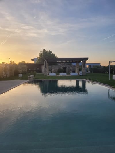 The image presents a tranquil outdoor scene at what appears to be a resort or private estate in Sogliano Cavour, Italy, captured during either sunrise or sunset. The sky exhibits a striking gradient, transitioning from a warm golden-yellow and orange hue near the horizon to a cool, light blue overhead, punctuated by wispy clouds and faint contrails.

In the foreground, a large infinity-edge swimming pool dominates, its calm, turquoise-blue water reflecting the sky and the surrounding architecture like a mirror. The pool's surface shows subtle ripples from a gentle breeze. Beyond the pool's concrete edge, a meticulously manicured green lawn extends towards a series of buildings. The primary structure visible is a modern-style building incorporating elements of stone masonry and dark, possibly wooden or metal, architectural beams. Several white, contemporary outdoor lounge chairs or daybeds are strategically placed on the lawn near the building. Trees with varying foliage are scattered across the landscape, providing natural accents.

No people are visible, contributing to the serene and undisturbed atmosphere. The low sun creates warm, ambient lighting across the scene, particularly highlighting the reflections in the water. No visible text is present.