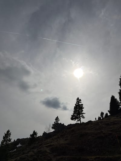 The image shows a landscape in Kamnik, Slovenia. The sky is mostly overcast with bright sunlight peeking through the clouds. A contrail from an aircraft is visible in the lower-left part of the sky. A cliff face with evergreen trees growing on it is on the right, oriented vertically.