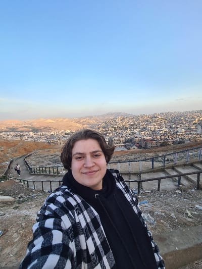 The image displays a young adult individual captured in a selfie on a hillside overlooking the city of Qudsaiya, Syria. The individual, positioned centrally and smiling faintly, has medium-length brown hair and is wearing a black hoodie underneath a black-and-white plaid flannel jacket. Behind the subject, a winding dirt path with wooden railings descends the hillside. Another individual is visible walking down this path in the background, to the left of the main subject. The terrain of the hillside is composed of dry, reddish-brown earth. In the mid-ground and far background, a dense urban landscape of buildings extends across the horizon, bordered by arid, light-brown hills. The sky is clear and light blue, indicating daylight conditions.