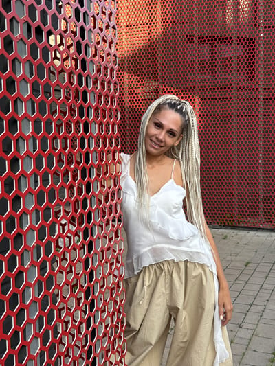 A woman with blonde braided hair stands next to a red hexagonal metal fence in Есбол ауылдық округі, Kazakhstan. She is wearing a white ruffled tank top and loose beige pants. The fence is a prominent feature in the foreground, with a hexagonal pattern. Behind the woman, a red industrial-style wall with a similar hexagonal pattern is visible. The ground is paved with grey concrete slabs and has patches of green grass. The scene appears to be outdoors in an urban or industrial setting.