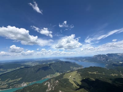 This is a high-angle, wide-shot, aerial photograph of a mountainous landscape with a body of water, likely a lake, in Sankt Gilgen, Austria. The sky is a vibrant blue with scattered white cumulus clouds. The foreground consists of steep, tree-covered slopes with visible hiking trails. In the middle ground, the emerald-green lake stretches out, bordered by smaller green hills and some structures that appear to be buildings or a small town. Further in the distance, larger, rugged mountains rise, their peaks partially obscured by haze. The overall scene depicts a serene natural environment on a clear day. There are no people or animals visible in the image.