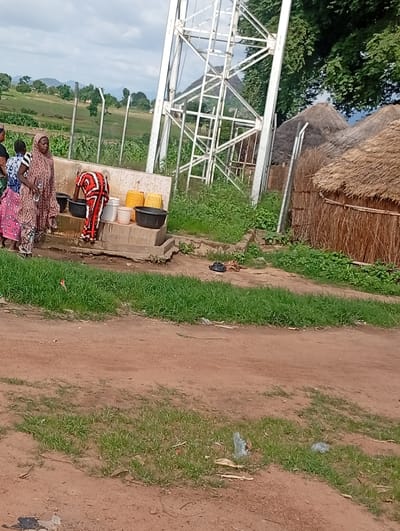 This image depicts a scene in Wuro Sani, Nigeria, near a water access point. Several individuals are present, with three women standing near a concrete structure that appears to be a communal water source or well. One woman, wearing a red and white patterned garment, is bent over, interacting with the water. Two other women stand to her left, one holding a plastic bottle. In front of the water source, there are at least three large containers: two yellow jerrycans and two black basins. Behind the water structure, the landscape transitions to rural settings with thatched-roof huts and vegetation. A tall metal tower, possibly a telecommunications tower or part of a water system, is visible in the background, towering over the scene. The ground in the foreground is a reddish-brown dirt path with patches of green grass. The sky is overcast with clouds.