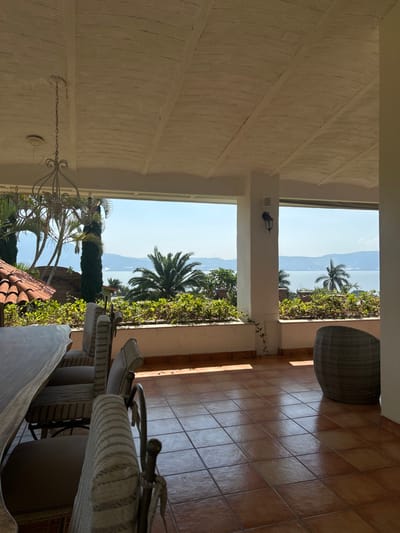 The image depicts a covered outdoor patio or terrace in San Juan Cosalá, Mexico, offering a panoramic view of a large body of water and distant hills under a clear blue sky. The patio floor is covered with terracotta-colored square tiles, reflecting strong sunlight that creates distinct shadows on the right side. The ceiling is light-colored, possibly off-white, featuring an arched or vaulted design with visible structural beams and a textured finish. A multi-arm, ornate white metal chandelier hangs from the ceiling on the left.

In the foreground and mid-ground, a long, light-colored wooden table with an irregular edge is positioned on the left, flanked by several chairs with metal frames and striped fabric cushions. A single dark-colored wicker-style armchair is situated on the right. White rectangular pillars support the roof structure. A low, white wall or planter box, filled with dense green foliage, extends across the width of the patio, defining the open viewing area. A black wall sconce is affixed to the central pillar.

Beyond the patio, the environment includes multiple palm trees and other green trees. A large expanse of calm, blue water, likely Lake Chapala, dominates the mid-ground, with a line of distant hills or mountains visible on the horizon. A section of a terracotta-tiled roof is visible on the far left, below the patio level. No people are present, and no specific actions or interactions are depicted. The scene is illuminated by bright daylight.