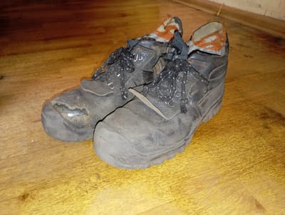 A pair of worn, black, leather work boots with orange and tan linings are placed on a light brown wooden floor. The boots are laced with black laces and show significant signs of wear, including scuffs and a tear on the toe of the left boot. The scene appears to be indoors, possibly a room in a residential building in Brocēni, Latvia, given the visible wooden flooring and baseboard. There are no people or active interactions depicted. The lighting suggests an artificial light source.