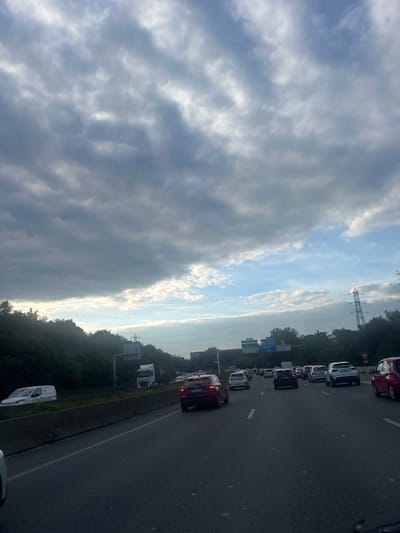 The image depicts a congested multi-lane highway, likely a section of a major route in or near Villeneuve-la-Garenne, France. The scene is observed from within a vehicle, facing forward in the direction of traffic.
The sky occupies the upper two-thirds of the frame, characterized by extensive, dense grey cloud cover, predominantly stratocumulus or altocumulus, with brighter patches and streaks of blue sky visible towards the right and along the lower horizon.
To the left and right of the highway, dense green foliage, consistent with trees and bushes, forms an embankment. On the right side, a tall metal lattice-work electricity pylon is visible above the tree line. In the distance, above the foliage on the right, blue highway signs with white text are partially discernible.
The highway consists of at least three lanes of traffic moving away from the viewer, marked by white dashed lines on a dark asphalt surface. A concrete barrier separates these lanes from the left shoulder or opposite direction of traffic, where a white van (possibly a Renault Kangoo or similar) is parked or stopped. Further down on this side, a white-cabbed semi-truck is also visible.
Multiple vehicles occupy all visible lanes, appearing either stationary or moving at a very slow pace, indicating heavy traffic congestion. These vehicles primarily consist of passenger cars of various colors (dark, white, silver) and sizes, with a dark SUV (possibly an Audi or similar model) with illuminated taillights prominent in the midground, slightly to the left of the center. A red hatchback is partially visible on the far right edge of the frame. No individuals are visible outside of vehicles. The scene is illuminated by natural daylight, with diffuse light conditions due to the cloud cover.